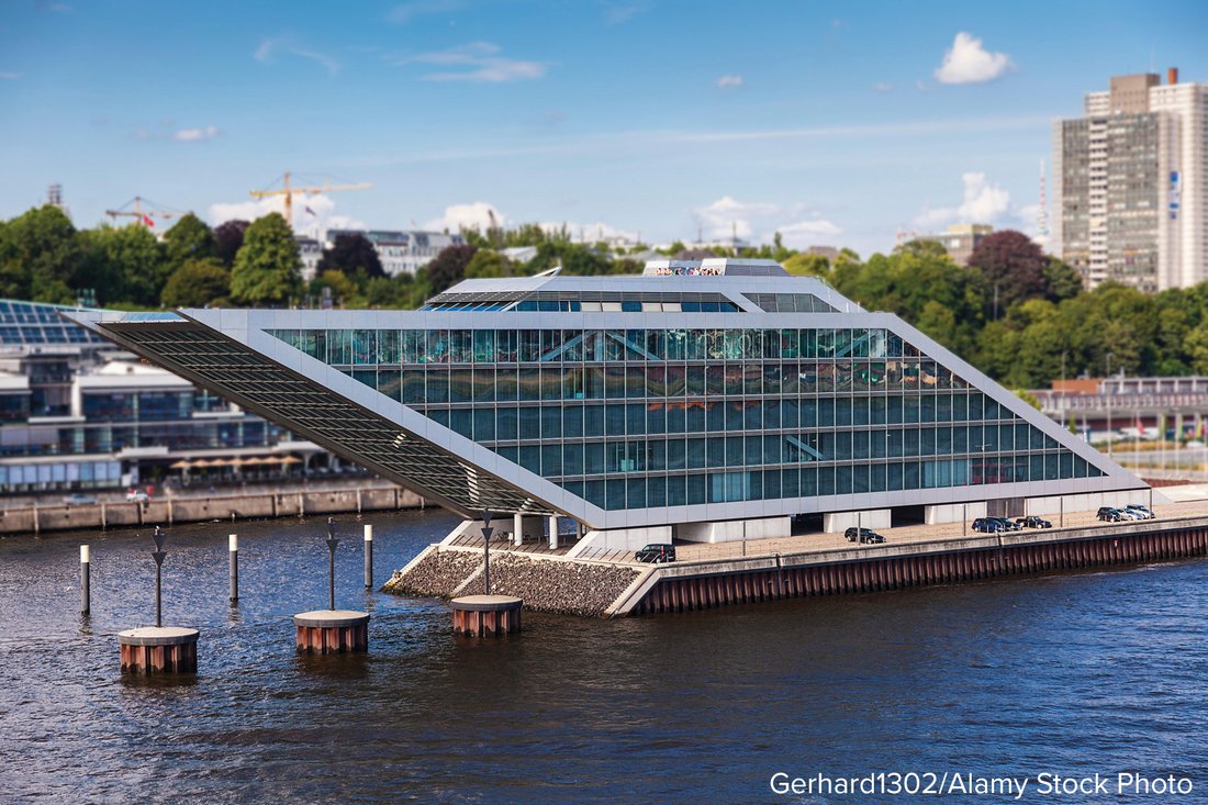 A photograph of a modern glass building with a sloping roof beside water.